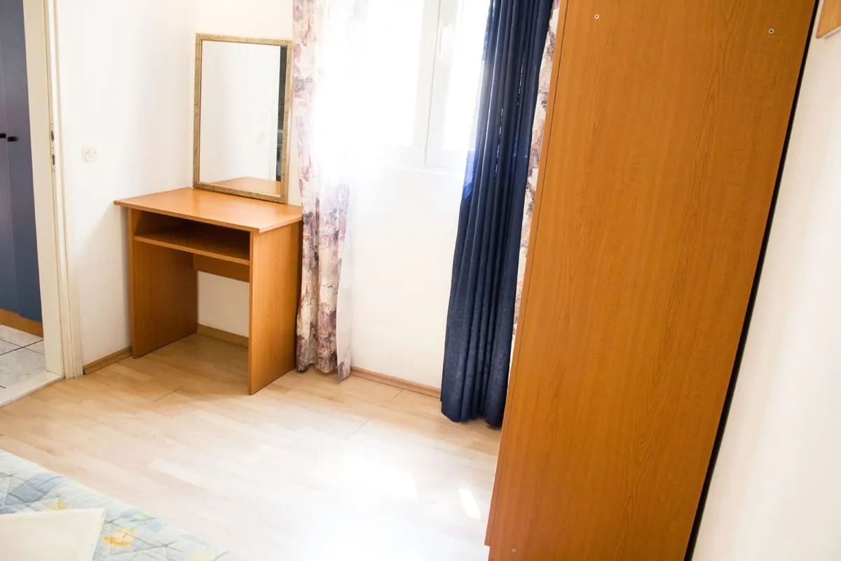 Bedroom corner with a desk, a mirror above it, and a tall wardrobe beside the window