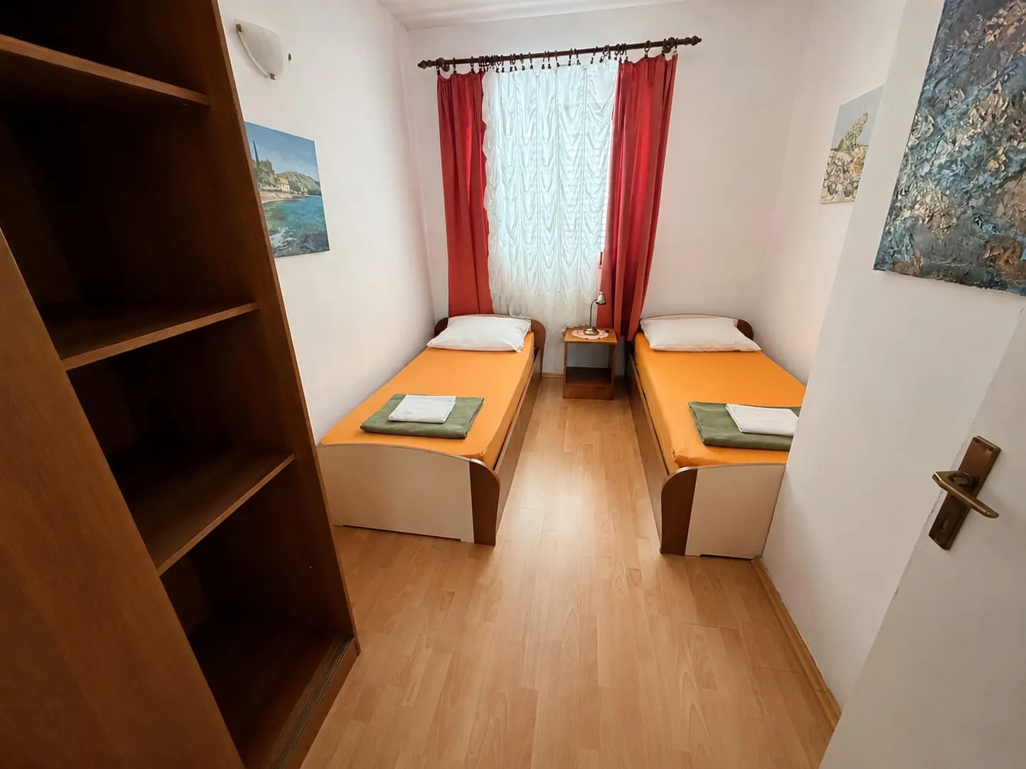 Two single beds with orange covers, a shelf unit, and red curtains at the window